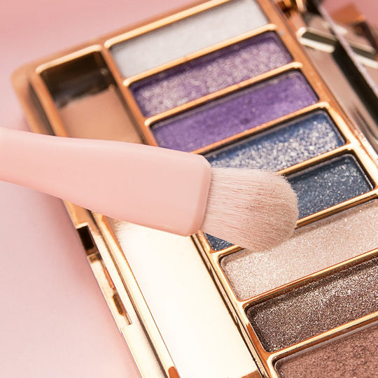 A pink makeup brush applying shimmering eyeshadow from a multi-color palette.