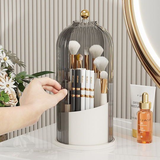 Hand reaching for makeup brushes inside a clear acrylic dome organizer on a vanity table.